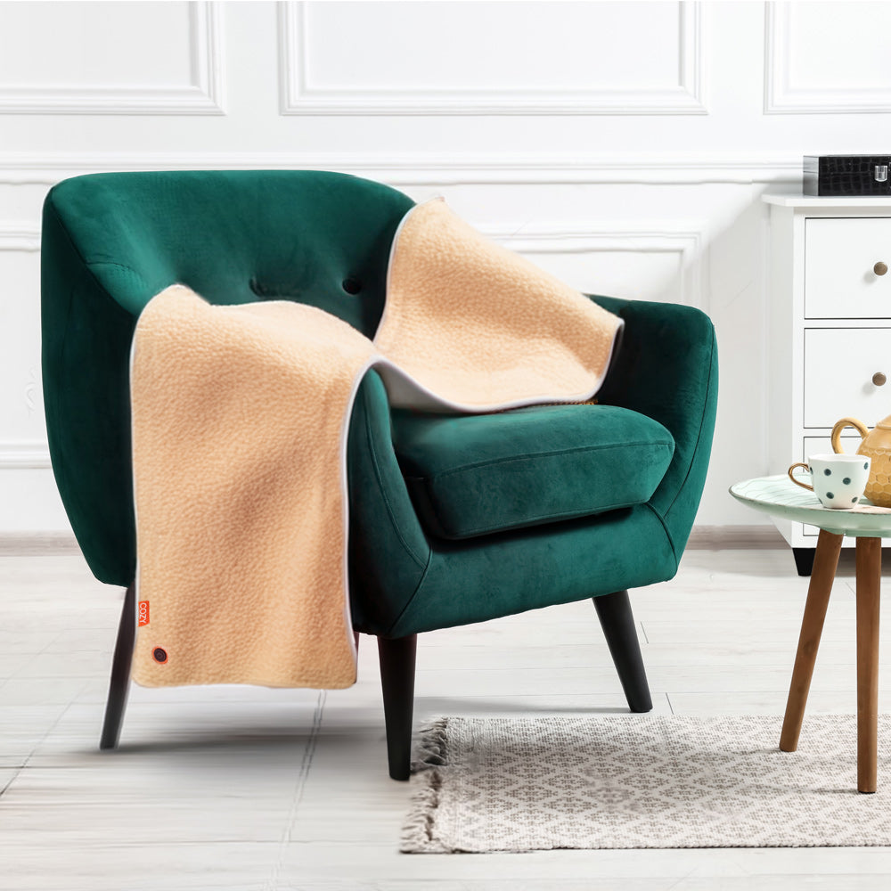 The COZY Heated Throw & Seat Cover Pad - JORV in beige is draped over a green velvet armchair beside a round table with tea and a cabinet, all set in a bright, modern room with white walls and a gray rug.