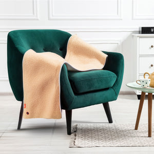 The COZY Heated Throw & Seat Cover Pad - JORV in beige is draped over a green velvet armchair beside a round table with tea and a cabinet, all set in a bright, modern room with white walls and a gray rug.