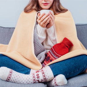 A person sits cross-legged on a couch, wrapped in the COZY Heated Throw & Seat Cover Pad - JORV, holding a mug. They wear a sweater, blue jeans, patterned socks, and have a red hot water bottle on their lap.