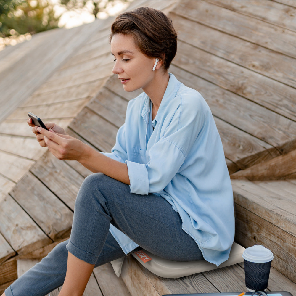 A woman with short hair, wearing wireless earbuds and a light blue shirt, sits on wooden steps. She looks at her smartphone, coffee beside her and enjoying extra comfort from the COZY Cordless Heated Seat Pad - JORD.
