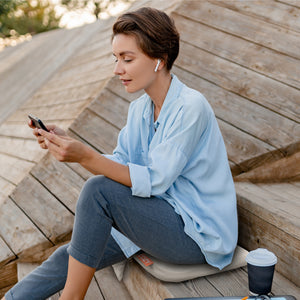 A woman with short hair, wearing wireless earbuds and a light blue shirt, sits on wooden steps. She looks at her smartphone, coffee beside her and enjoying extra comfort from the COZY Cordless Heated Seat Pad - JORD.