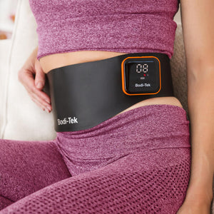 A person in a pink workout outfit wears the Ab & Back Muscle Strengthener with heat, which features a digital screen displaying numbers and has a sleek black and orange design.