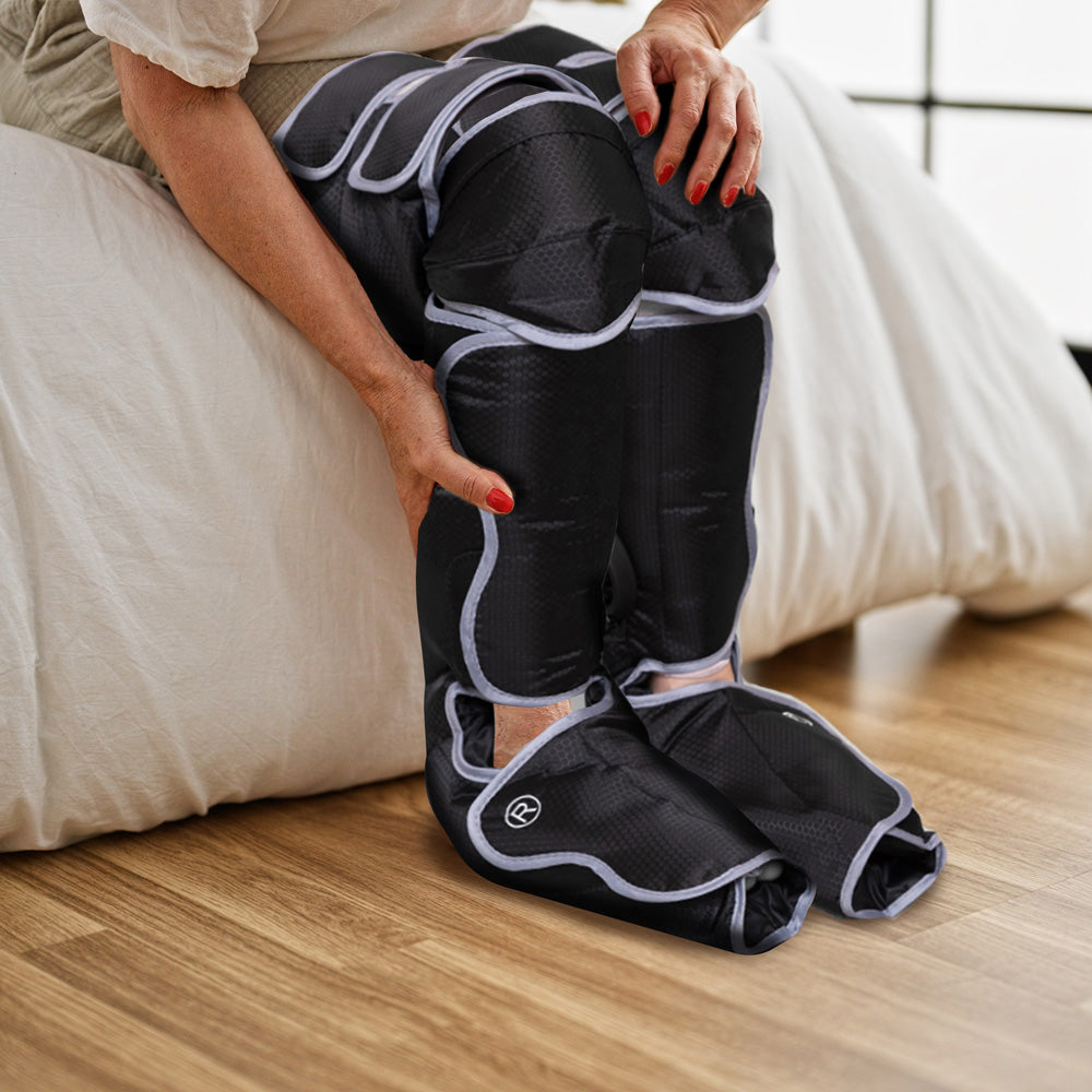 A person on a bed wears the Comfort360° Air Compression Full Leg Massager Boot, which covers their feet, calves, and thighs for massage or pain relief. The room features wooden floors and a window in the background.