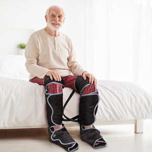 An elderly man with a white beard sits on a bed in red plaid pajama pants, using the Comfort360° Air Compression Half Leg Massager Boot for lower leg therapy. The room is bright with white bedding and natural light.