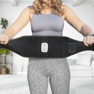 A woman in athletic wear holds a Smart Fitness EMS Muscle Toning Belt with a control button, standing in a bright, modern living room—ready to enhance her abs firming routine.