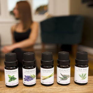 Five bottles from the Rio Aromatherapy 100% Essential Oil Collection—Peppermint, Lavender, Lemongrass, Eucalyptus, and Tea Tree—are lined up on a table. A woman sits in the blurred background, looking relaxed.