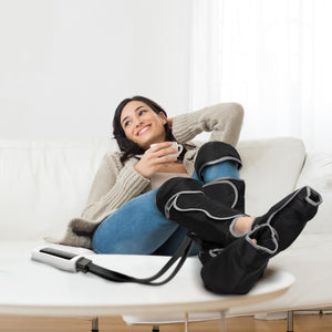 A woman lounges on a white sofa in a beige sweater and blue jeans, smiling and holding a cup while using the Comfort360° Air Compression Half Leg Massager Boot with multiple massage programs for cozy relaxation.