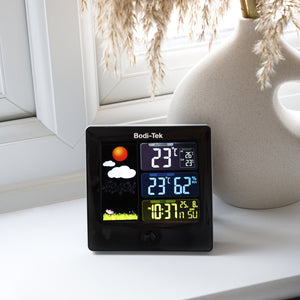 A Digital Weather Station rests on a white windowsill next to a beige vase with pampas grass, displaying temperature (23°C), humidity (62%), time, date, and weather icons on its wireless monitor.