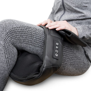 A person in a gray tracksuit uses the Neck & Shoulder Shiatsu Massager with control buttons on their thigh. The black device provides deep kneading muscle relief while their hand rests on it.