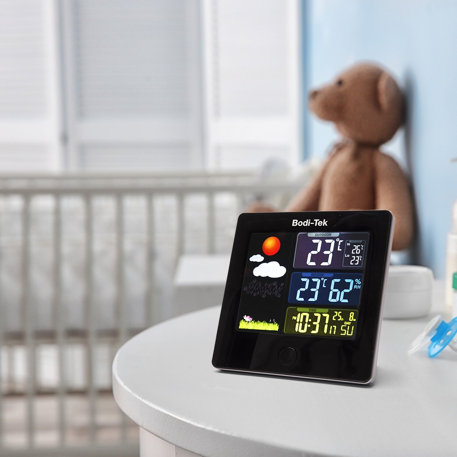 A Digital Weather Station rests on a white nursery table, displaying indoor/outdoor temperature, humidity, and time. A crib and brown teddy bear appear in the softly lit background.