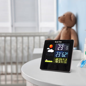 A Digital Weather Station rests on a white nursery table, displaying indoor/outdoor temperature, humidity, and time. A crib and brown teddy bear appear in the softly lit background.