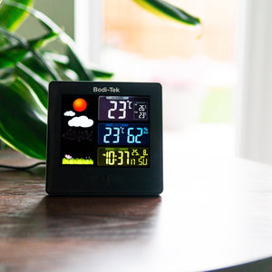 The Digital Weather Station sits on a wooden table, showing indoor and outdoor temperature, humidity, time, and a weather icon. A green plant and sunny window are blurred in the background.
