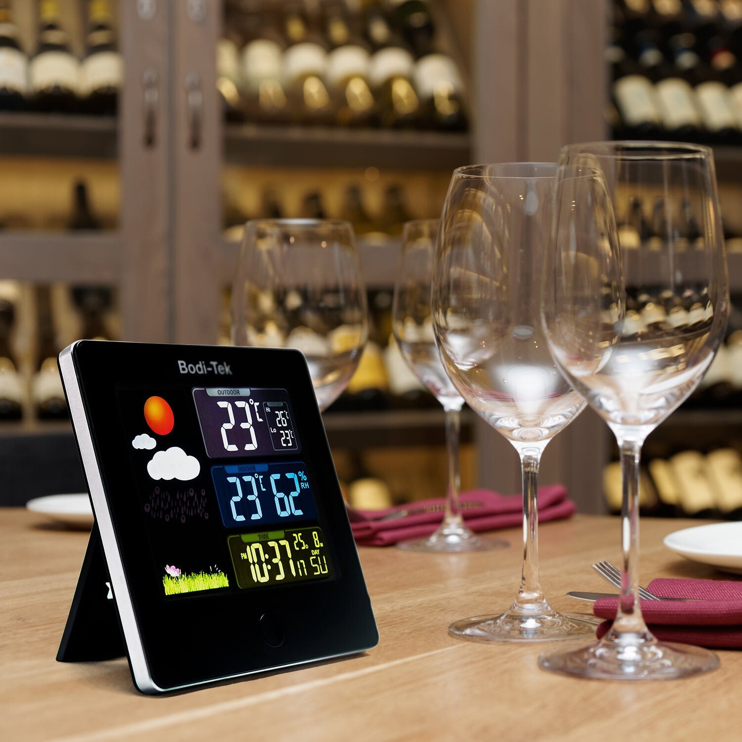 A Digital Weather Station sits on a set dining table with wine glasses in the foreground and a wine rack filled with bottles visible in the background.
