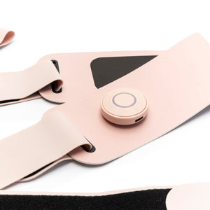 Close-up of the Ab, Tone & Shape EMS abdominal belt in light pink with adjustable straps, a circular control button, and a black rectangular section for muscle stimulation, shown worn on the body against a white background.