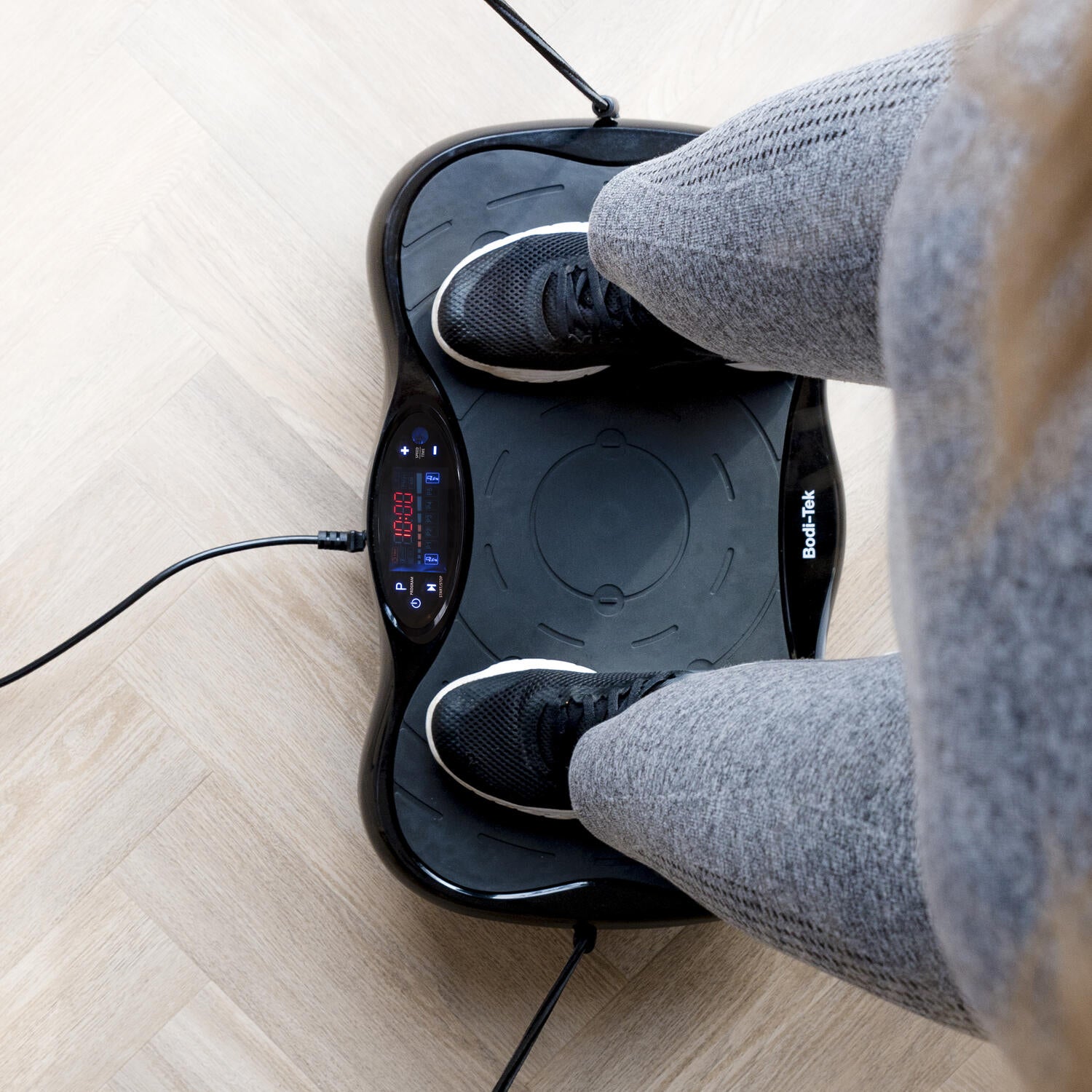 A person stands on the Whole Body Vibration Training Exercise Plate with a visible display, wearing gray leggings and black sneakers, ready for a full body workout using vibration technology on a light wooden floor.