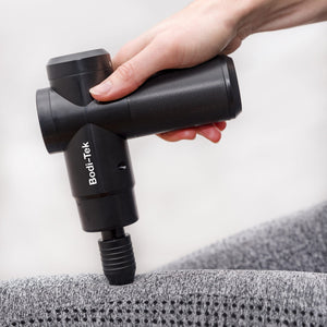 A hand holds a black Hot & Cold Deep Tissue Massager Gun, pressing it against a grey textured fabric surface.