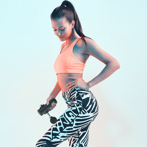 A woman in a peach sports bra and patterned leggings uses the Mini Deep Tissue Massage Gun on her thigh while standing against a light blue background.