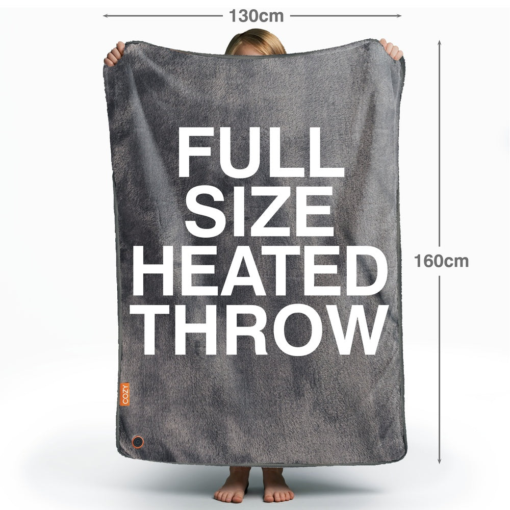 A person holds a large grey Cozy Wireless Rechargeable Snuggle Blanket Heated Throw (130 x 160 cm) with bold white text saying FULL SIZE HEATED THROW; only their feet, hands, and part of the head can be seen.