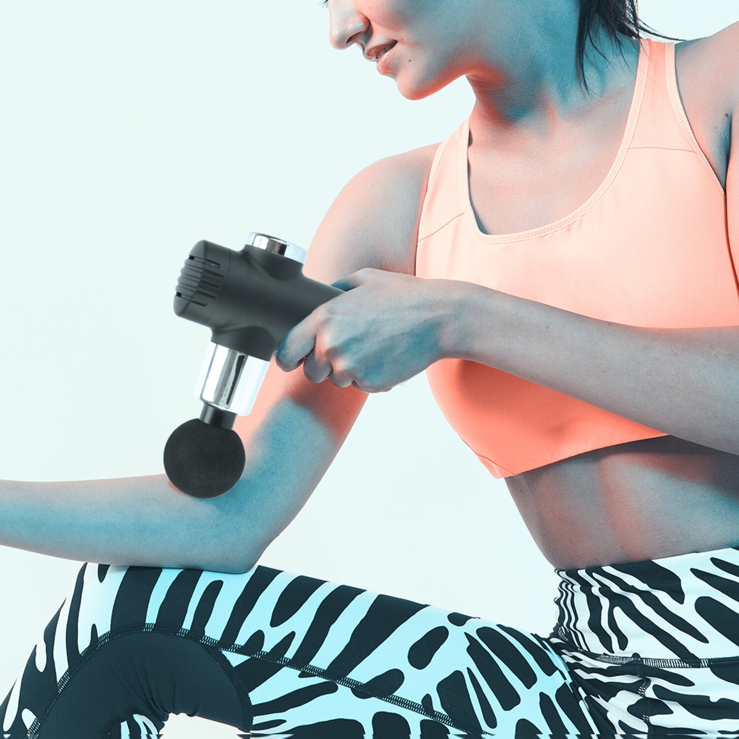 A woman wearing a peach sports bra and zebra leggings sits against a light blue background, using the Mini Deep Tissue Massage Gun on her forearm for muscle relief.