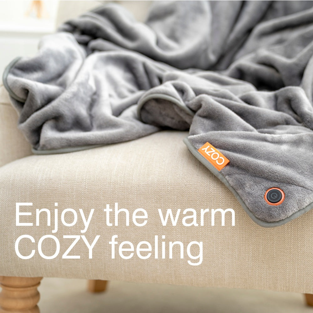 A soft gray Cozy Wireless Rechargeable Snuggle Blanket Heated Throw is draped over a beige armchair. Large white text on the image reads, Enjoy the warm COZY feeling.