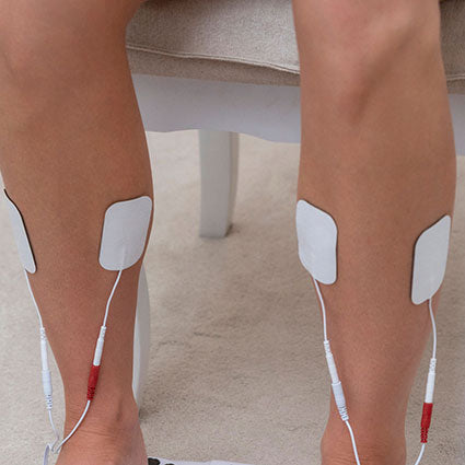 Close-up of a person’s lower legs with two pairs of Reviber Premium Electrode Gel Pads attached by wires—ideal for use with circulation boosters or electrical muscle stimulation devices. The person sits on a chair over a carpeted floor.