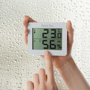 A person holds a Digital Room Thermometer and Hygrometer showing 23°C and 56% humidity, pointing to its screen, while water droplets are visible on the window in the background.