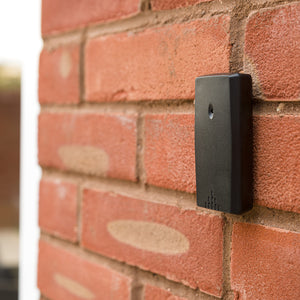 A Digital Weather Station in black is mounted on a red brick wall, slightly angled, featuring a small hole and ventilation slits on its surface.