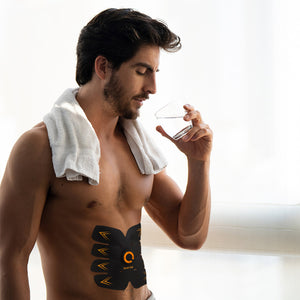 A shirtless man with a white towel around his neck drinks water, using the Ab Trainer EMS abdominal muscle trainer attached to his stomach. Sunlight from a window streams in behind him.