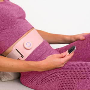 A person in a pink fitness outfit sits on a couch, wearing the Ab, Tone & Shape EMS abdominal belt around their waist and holding its small black remote control.