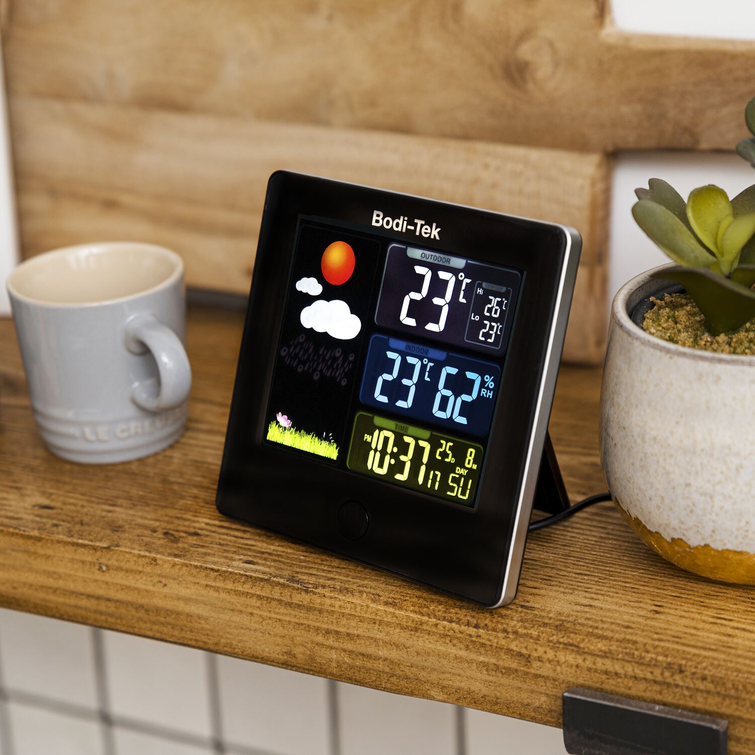 A Digital Weather Station rests on a wooden shelf beside a gray mug and a potted succulent, showing indoor/outdoor temperature, humidity, time, date, and a weather graphic of sun and clouds.