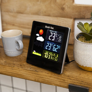 A Digital Weather Station rests on a wooden shelf beside a gray mug and a potted succulent, showing indoor/outdoor temperature, humidity, time, date, and a weather graphic of sun and clouds.