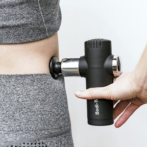 Wearing gray athletic clothing, a person uses the Mini Deep Tissue Massage Gun, a black percussive device, on their lower back.