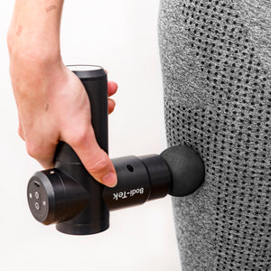 A person uses a black Hot & Cold Deep Tissue Massager Gun on their thigh, wearing grey patterned athletic pants. The massager is applied to aid muscle recovery and deliver soothing relief.