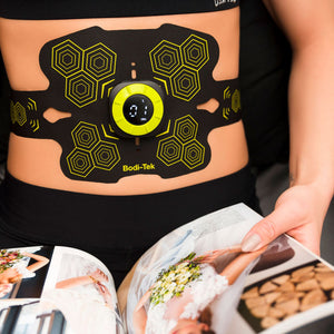 Wearing the Abs and Oblique Trainer Pro with a digital display, a person browses a bridal magazine of wedding dresses, working on core ab muscles for their big day.