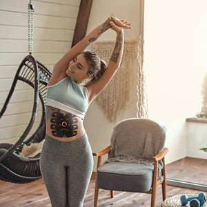 A tattooed woman stretches in a cozy, sunlit room, wearing a crop top and leggings that show off her results from using the Ab Trainer. A hanging chair, armchair, and stylish decor enhance the inviting background.