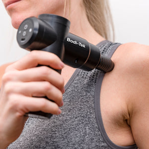 A person uses the Hot & Cold Deep Tissue Massager Gun in black on their upper shoulder near the collarbone while wearing a grey athletic tank top, targeting the area for effective muscle recovery.