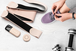 A person ties the laces of a purple sneaker near fitness gear, including dumbbells, resistance bands, the Ab, Tone & Shape EMS abdominal belt, a wearable device, and a remote control on a light wooden surface.
