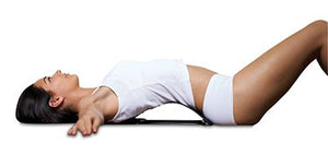 A woman in white athletic wear lies on her back, arms outstretched, using The Original BackMagic with Thermal Gel Heat Pads for lower back support and pain relief against a plain white background.