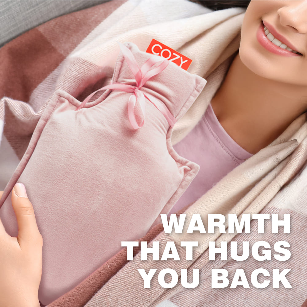 Wrapped in a blanket, a smiling person in a light pink top holds the Cozy UNA Rechargeable Hot Water Bottle. The bottle is soft pink and labeled "COZY," with text reading: Warmth that hugs you back.