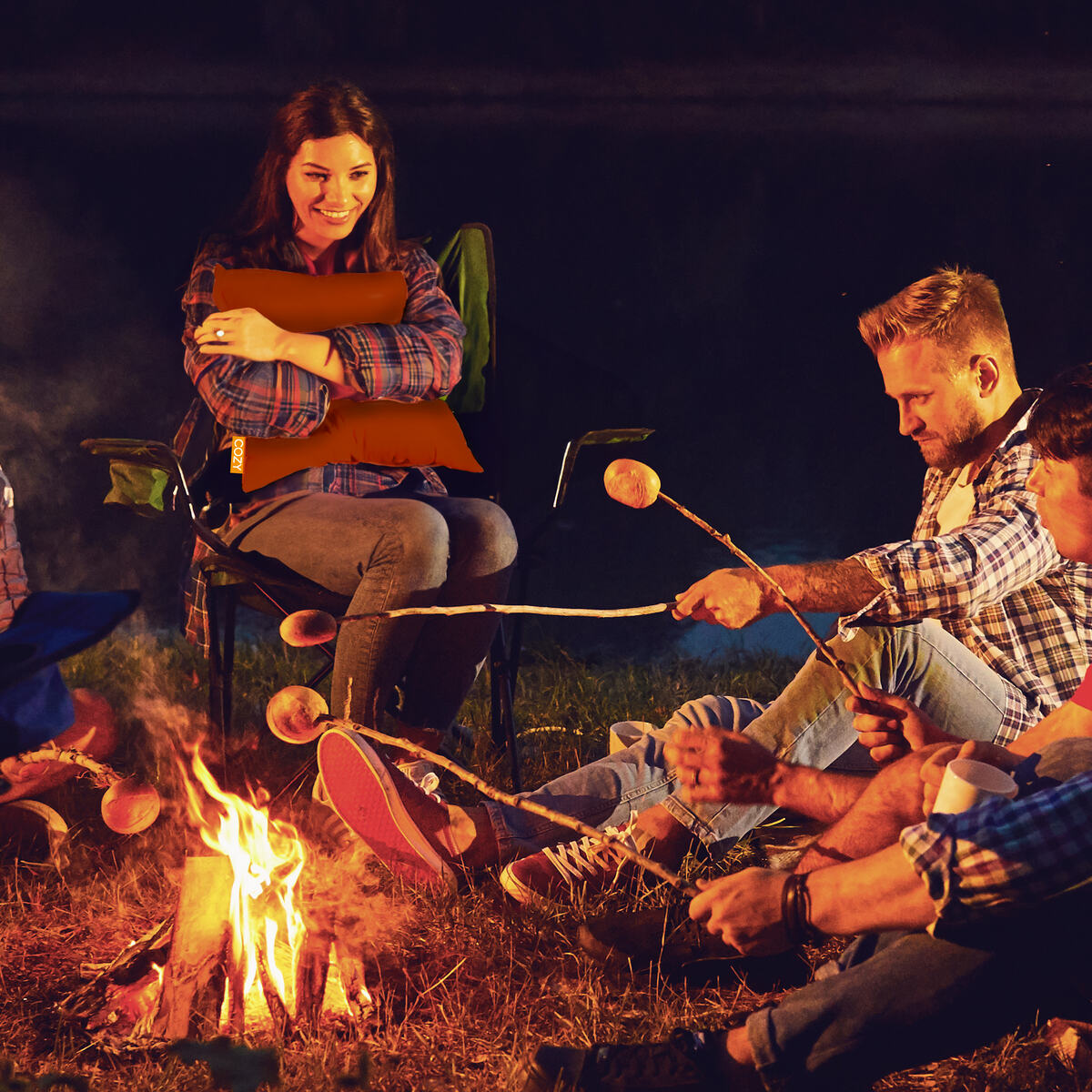 Group of friends sat around a fire - young female sat on a chair hugging heated orange cushion.