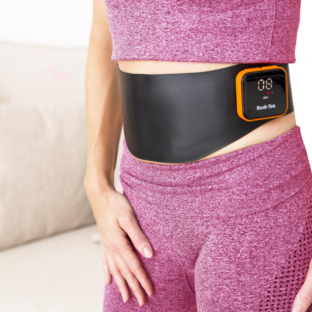 A person in a pink sports outfit wears the Ab & Back Muscle Strengthener with heat, featuring a small screen showing numbers, fastened around their waist.
