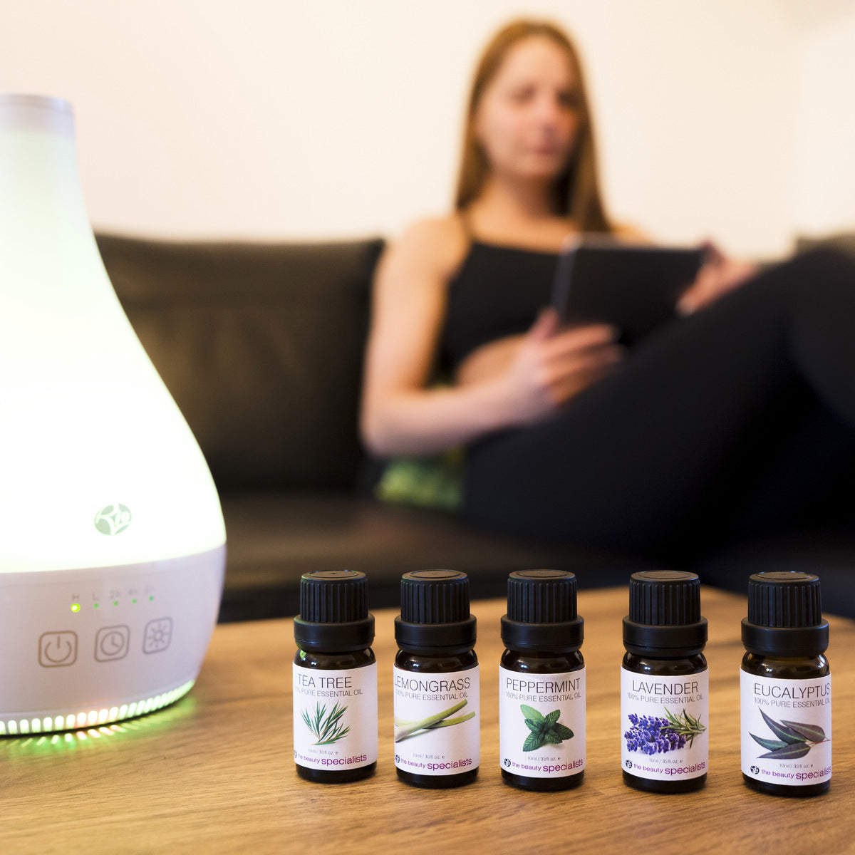 Five bottles of Rio Aromatherapy 100% Essential Oil Collection are lined up on a wooden table before a glowing diffuser, while a woman relaxes in the background with a tablet.