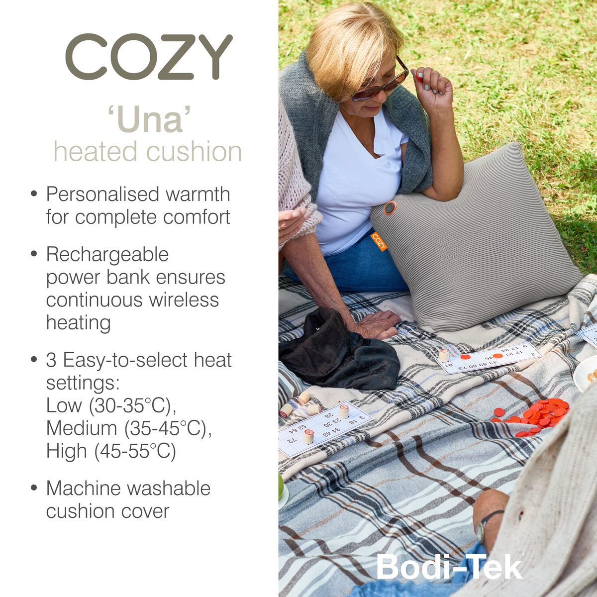 A woman sits on a picnic blanket with a grey ribbed COZY Heated Cushion Cordless - UNA Knitted (45cm x 45cm).