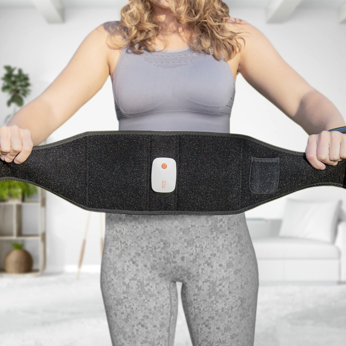 A woman in athletic wear holds a Smart Fitness EMS Muscle Toning Belt with a control button, standing in a bright, modern living room—ready to enhance her abs firming routine.
