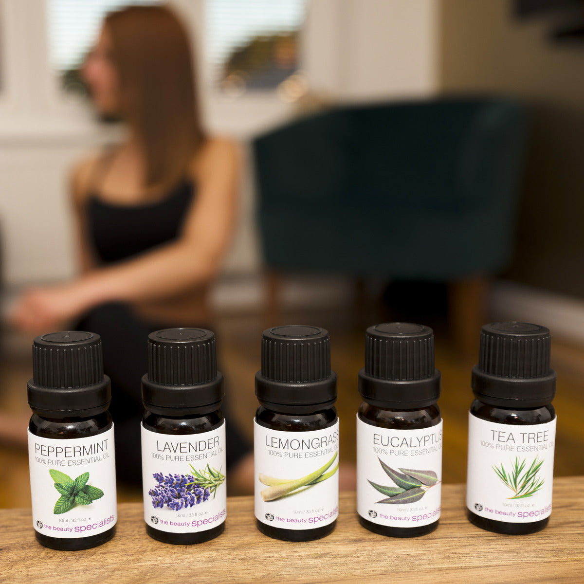 Five bottles from the Rio Aromatherapy 100% Essential Oil Collection—Peppermint, Lavender, Lemongrass, Eucalyptus, and Tea Tree—are lined up on a table. A woman sits in the blurred background, looking relaxed.