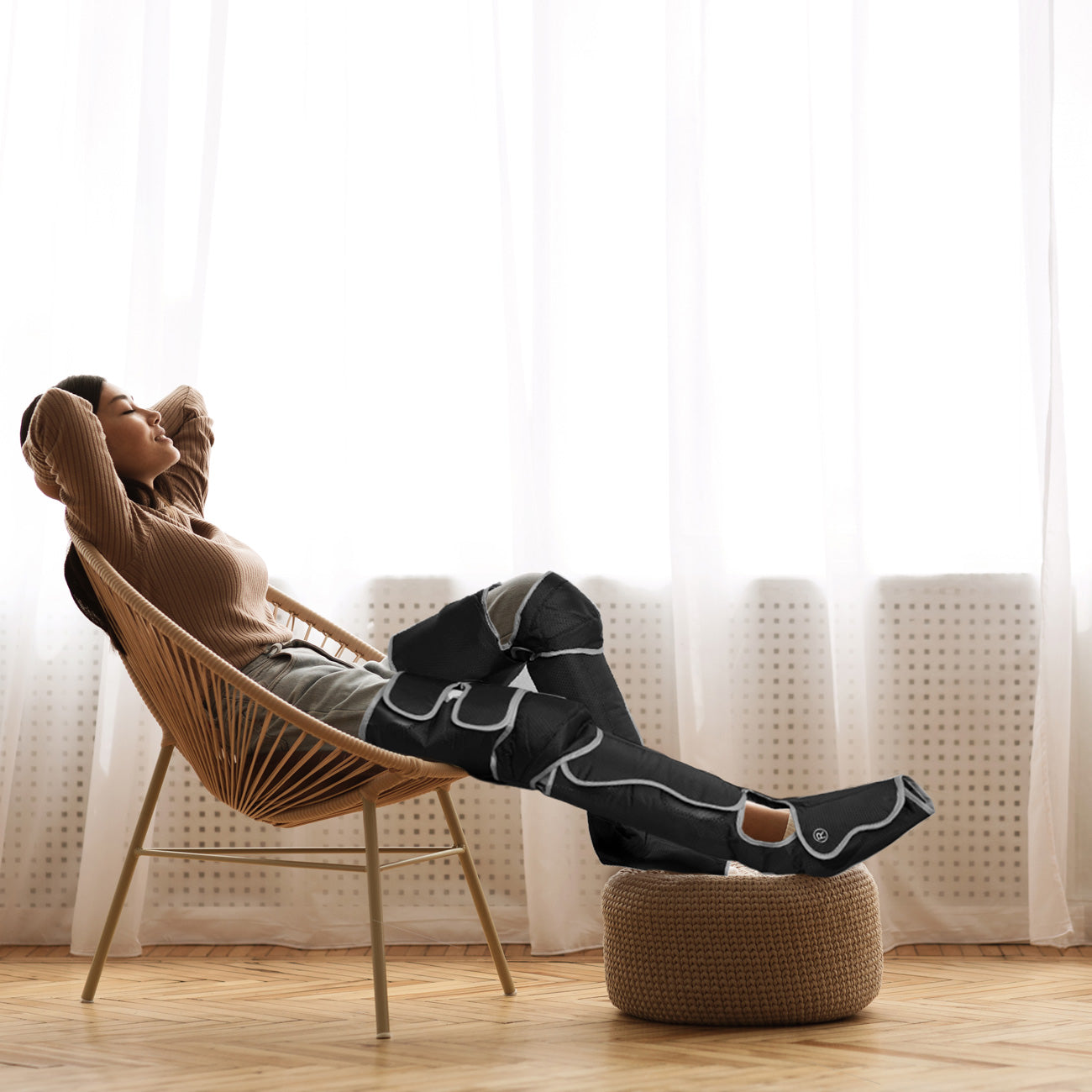 A woman relaxes in a chair with her legs elevated on a pouf, enjoying the soothing effects of the Comfort360° Air Compression Full Leg Massager Boot as light filters through sheer curtains, enhancing the cozy atmosphere.