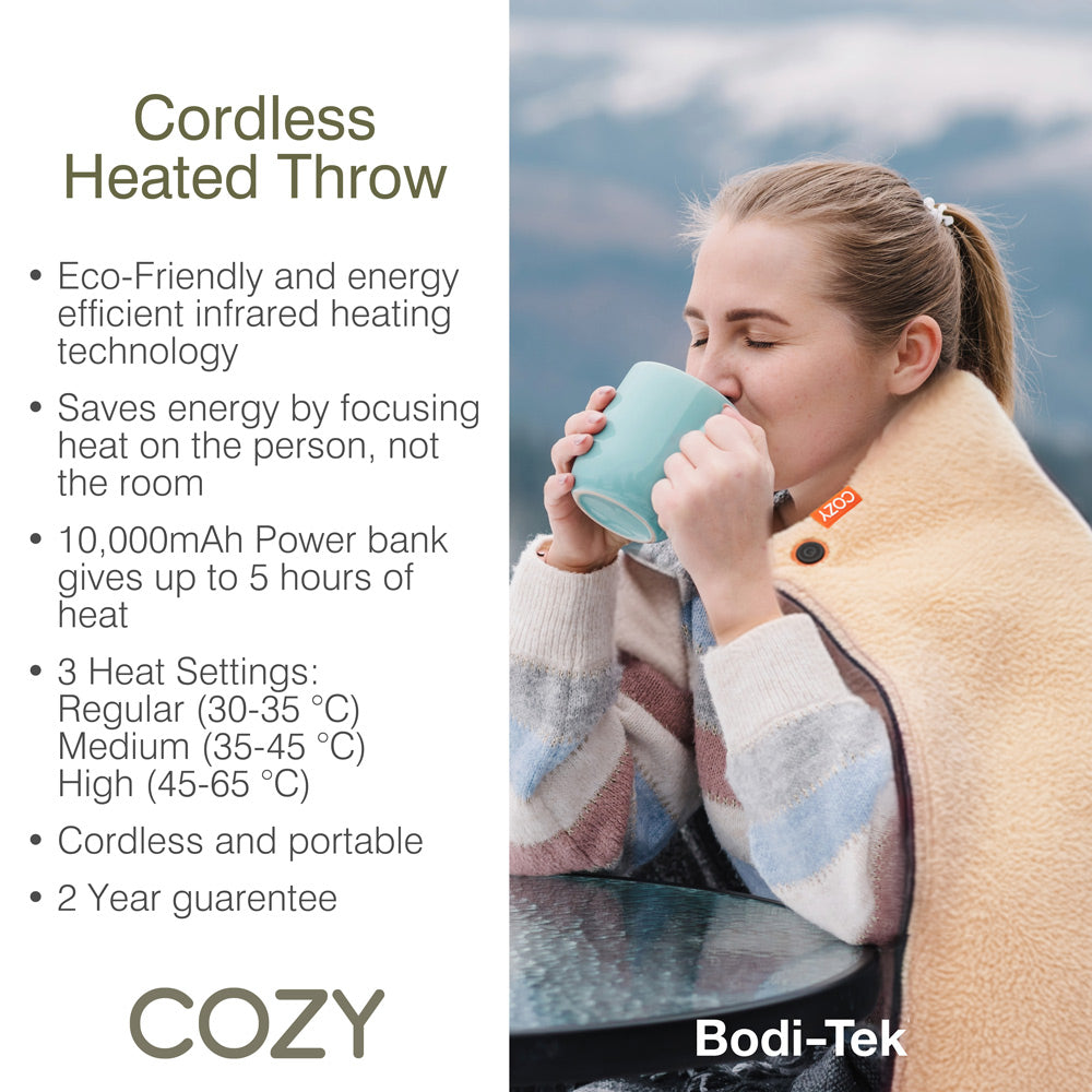 A woman sits outdoors wrapped in the tan COZY Heated Throw & Seat Cover Pad - JORV, sipping from a blue mug. Features include eco-friendly heating, power bank, 3 heat settings, portability, and a 2-year guarantee.