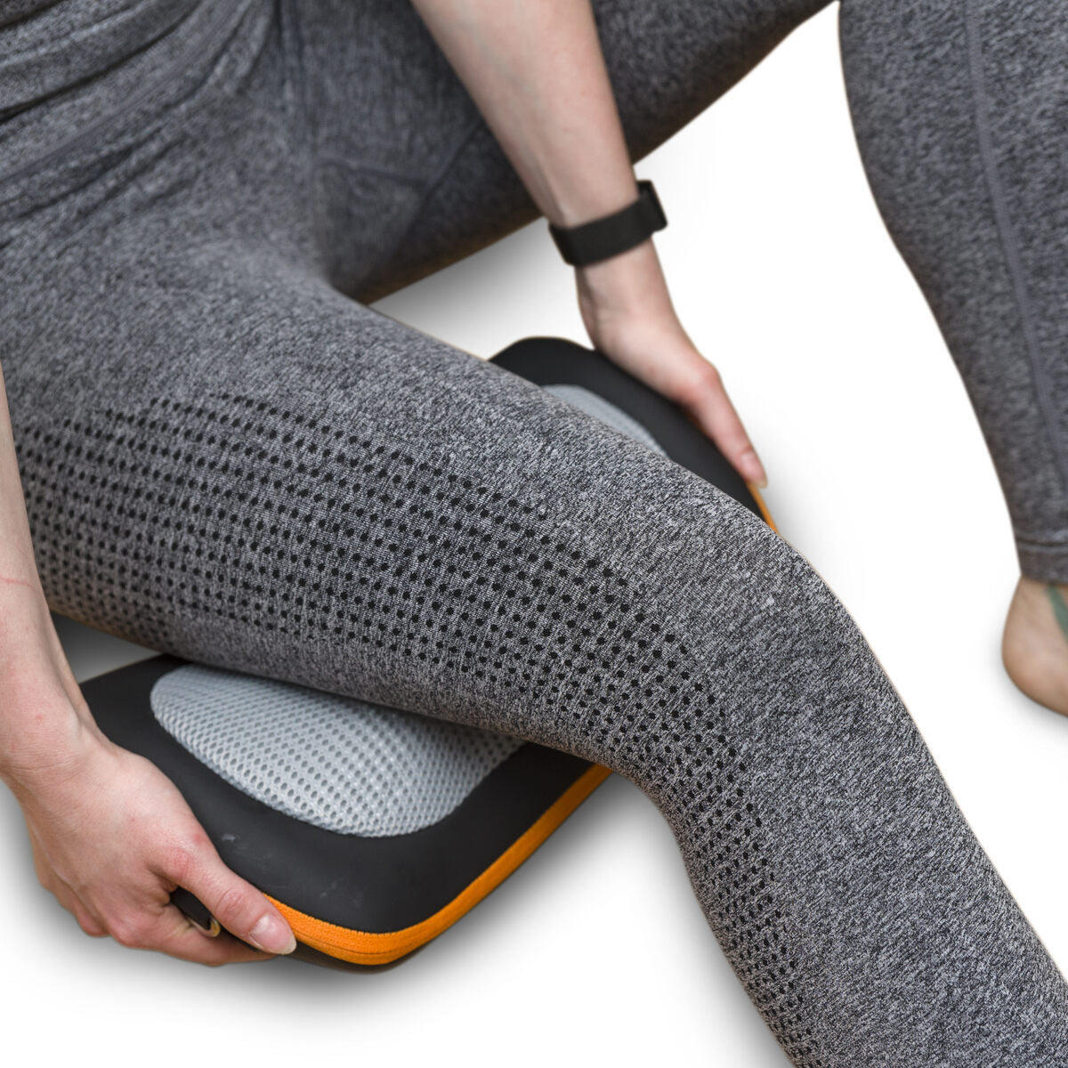 A person in gray athletic wear uses the Neck, Shoulder & Body Shiatsu Massager on their thigh while sitting on the floor, with only their legs, hands, and a fitness tracker-wearing arm visible.