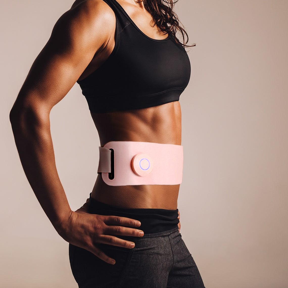 A fit woman in athletic wear stands sideways, highlighting the pink Ab, Tone & Shape EMS abdominal belt around her toned stomach, emphasizing its ab toning effect against a neutral background.