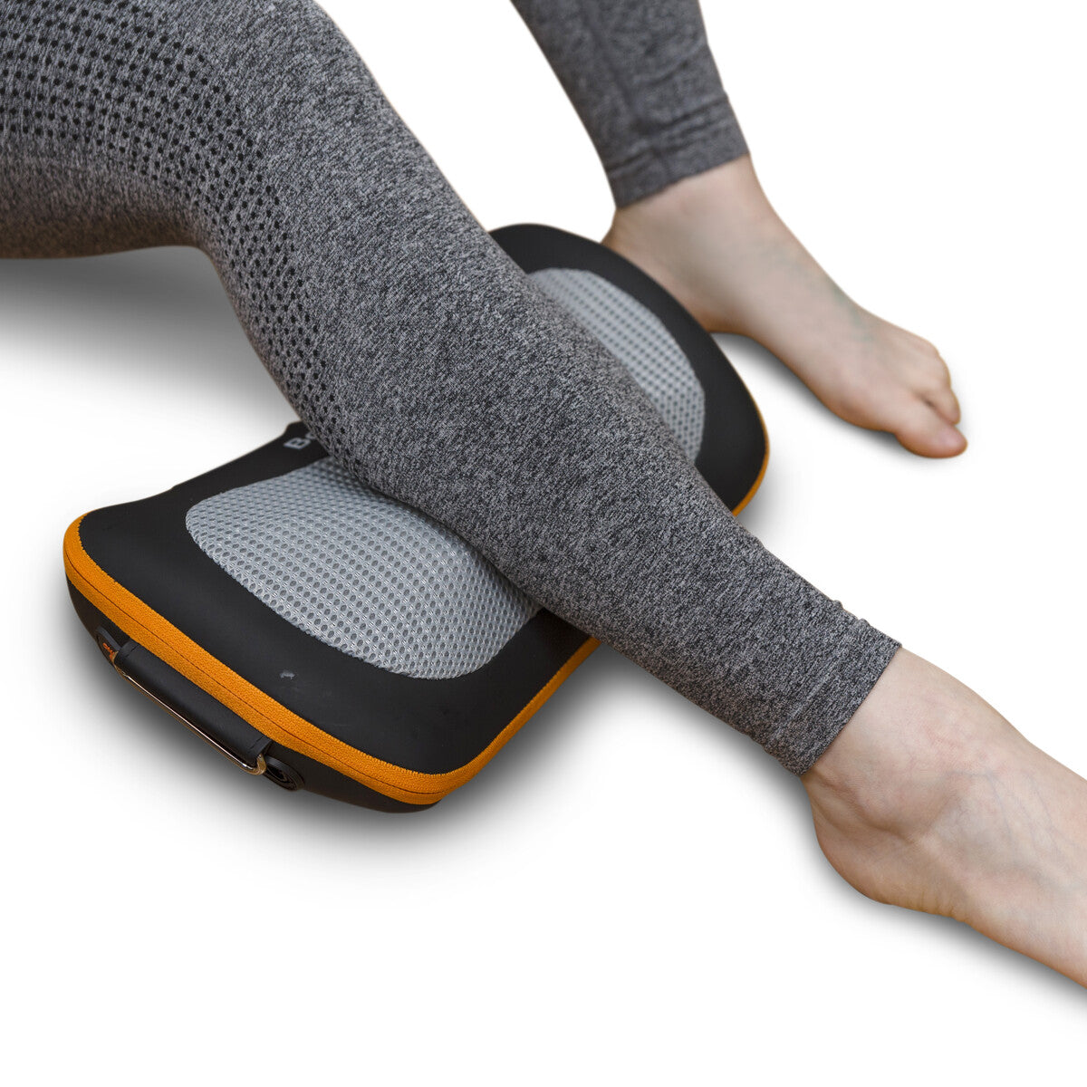 A person in gray leggings rests their legs on the Neck, Shoulder & Body Shiatsu Massager with black, gray, and orange accents, placed on a white surface.